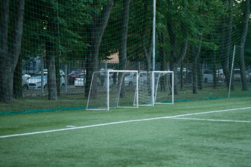 Two small soccer goals are positioned on a green field, framed by tall trees. The setting sun casts a warm light, highlighting nearby parked cars, suggesting a quiet afternoon