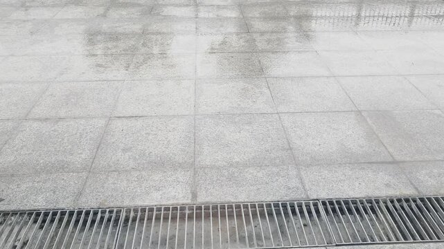 Rain-drenched tiled pavement with a horizontal metal drain grate and visible raindrop splashes.