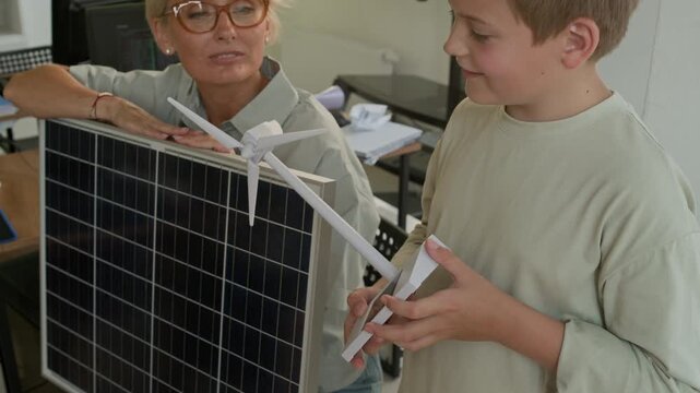 Cute schoolboy blowing on turbine of windmill mockup while teacher with sollar panel helping him during presentation of usage of renewable sources of energy