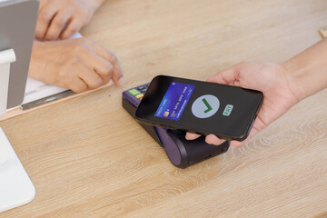 Hand holding smartphone making contactless payment with credit card via NFC on POS terminal at counter, illustrating modern cashless transaction convenience for digital financial technology use