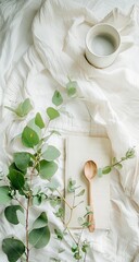 Soft, white draped fabric, green eucalyptus sprigs, a book, and a wooden spoon.  A cream-colored bowl sits atop the fabric