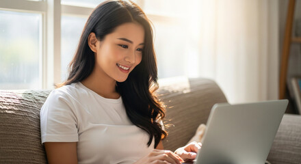Fototapeta premium A young chinese woman smiles brightly while typing on a laptop computer while sitting on a comfortable couch near a window during a sunny day.