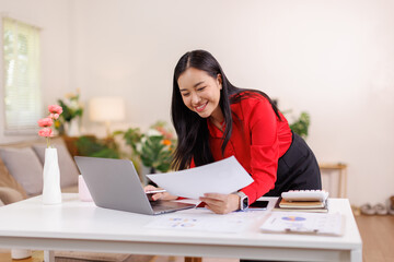 Portrait of a asian woman using laptop work on documents and a tablet while sitting on a desk at home
