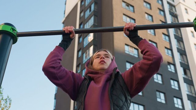 Sky view female exerciser working out intensively on pull up bar against urban building backdrop emphasizing forearm strength focus and control during outdoor calisthenics session