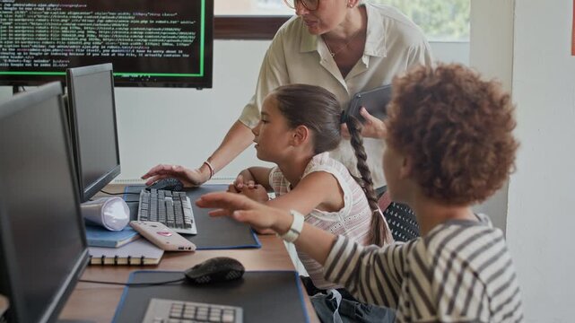 Cute schoolboy with curly hair sitting by desk in front of computer monitor and looking at diligent classmate while teacher of programming helping her at lesson