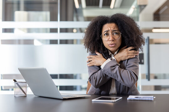 An African American young businesswoman sits at a desk in the office and is freezing from the cold, warming herself by hugging her arms