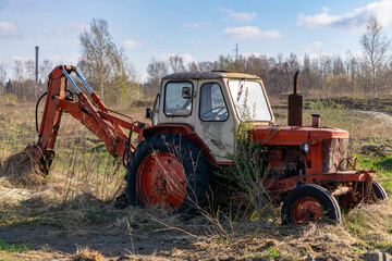 Old rusty tractor with backhoe abandoned in a field, damaged agricultural machine, partially overgrown with plants.