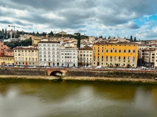 Colorful historic buildings along the Arno River with reflections in the water, framed by dramatic clouds above the cityscape.