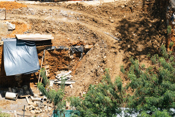A construction site in progress shows an uneven excavation with exposed earth, rocks, pipes, and buckets strewn around. A makeshift plastic tarp provides cover and green pine trees border the scene