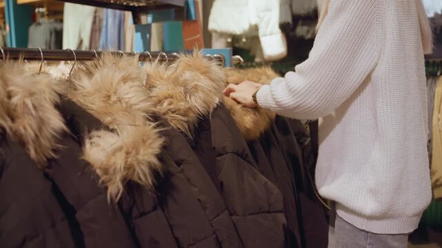 Trend setter browsing coat rack feeling fur trim assessing style and quality under warm boutique lighting with shoes displayed on top amid organized winter apparel racks evoking shopping ambience