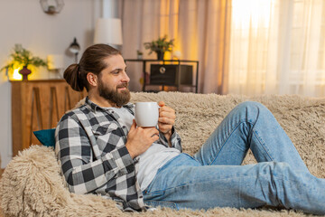 Young man resting lying at home, holding warm cup of coffee or tea with gentle grin and eased shoulders. Caucasian guy on sofa feels soothing quiet, appreciating short pause from busy tasks and noise.
