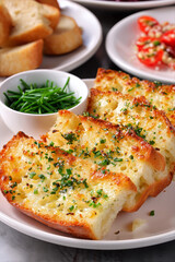 Golden garlic bread slices sprinkled with fresh chopped chives, served on a white plate with herbs and bread in background.