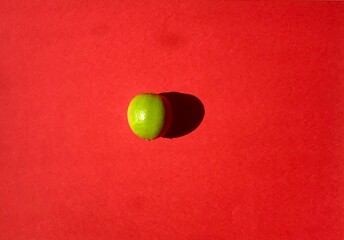 A solitary green lime is positioned centrally on a bright red background, illuminated by warm afternoon sunlight, emphasizing its vibrant color and smooth texture