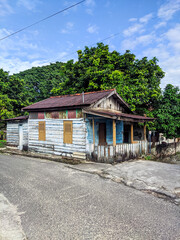 Traditional wooden house with tin roof stands by the roadside, showing signs of age and simplicity. Surrounded by lush green trees, it reflects rural charm and the heritage of local living.