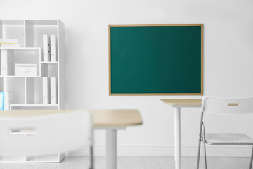 Blank green chalkboard, desks, chairs and shelving unit in classroom