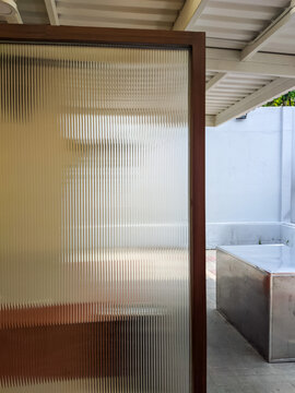 Frosted glass door with a wooden frame reveals a blurred silhouette, opening onto a minimalist outdoor patio with a concrete bench and corrugated roof in natural daylight.