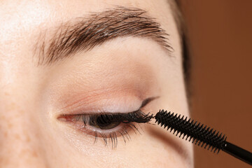 Woman applying mascara onto her eyelashes on brown background, closeup