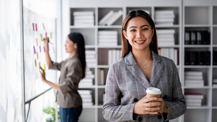 Young Adult Asian Businesswoman Smiling Holding Coffee Cup in Modern Office with Colleague...