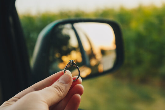 Woman holds engagement diamond ring in front of the side mirror with a view on the road in the meadow at sunset. Beautiful view from the car to the field. Travel concept, marriage proposal on holidays