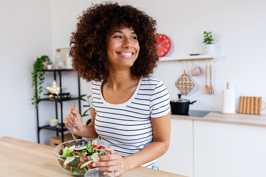 Young happy woman with curly hair smiling and eating healthy fresh salad in her modern kitchen at home. Healthy eating habits lifestyle and food concept