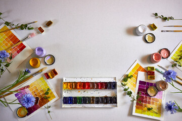 Flat lay of colorful watercolor palette, brushes, jars of paint, floral elements, and painted swatches on paper. Artistic workspace background, perfect for creativity, design projects, and inspiration