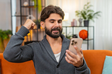 Indian man at home uses smartphone for mobile game win smiles reads message with news raises fist in joy celebrates victory. Arabian guy on sofa checks score happy thrilled does yes gesture win mood