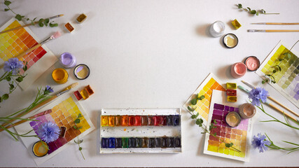 Flat lay of colorful watercolor palette, brushes, jars of paint, floral elements, and painted swatches on paper. Artistic workspace background, perfect for creativity, design projects, and inspiration