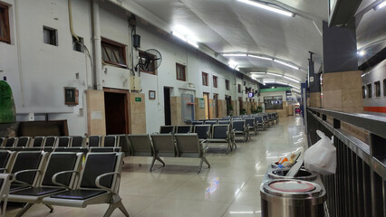Interior view of a train station waiting area with rows of empty metal chairs, bright ceiling lights, and a clean tiled floor. The long corridor design emphasizes order, waiting, and public transport 