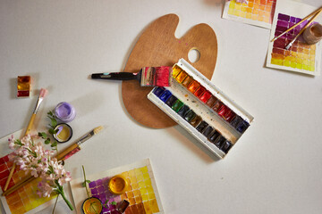 Artistic flat lay with watercolor palette, brushes, color swatches, flowers, and a wooden palette on a light background. Creative workspace with bright colors, perfect for design mockups