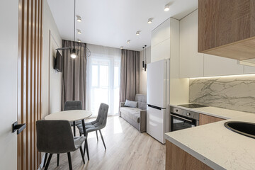 modern, compact studio apartment featuring a dining area, kitchen, and living space with a sofa near large windows. The color palette is neutral and contemporary