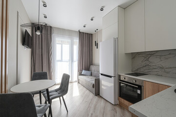 cozy modern apartment with a dining area, sofa, and a kitchen corner featuring white cabinets and a refrigerator. The room is bathed in natural light