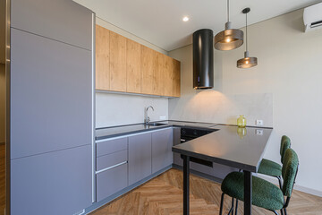 modern kitchen with gray and wood cabinets, a black range hood, pendant lights, a gray countertop with bar seating, and green chairs on a wooden herringbone floor