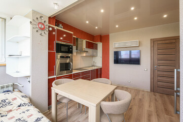 combined kitchen and living area in an apartment with modern design. kitchen area features red cabinets and stainless steel appliances. wooden dining table and chairs are placed near kitchen