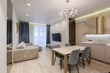 Bright, modern living space with a beige sofa, dining table, and sleek kitchen area. A unique bubble chandelier hangs above