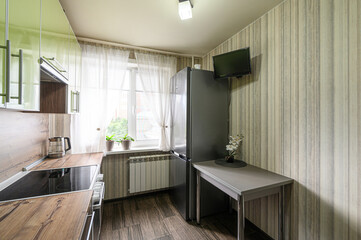 cozy kitchen with a refrigerator, a small table, green cabinets, a stove, and a window with sheer curtains. A TV is mounted on the striped wallpaper