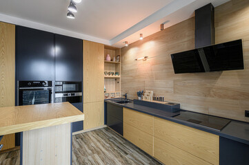Modern kitchen with wood accents, black cabinets, and a black countertop. Sleek appliances and stylish lighting complete the design