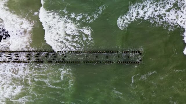 Aerial view of ocean waves crashing against rows of stone breakwaters. The geometric pattern of the structures contrasts with the dynamic movement of the sea, creating a striking coastal scene.