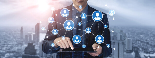 A business professional engages with a digital device, showcasing a virtual network of people. The backdrop of a modern urban skyline enhances the concept of connectivity. Tessel