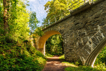 Old railway stone arch bridge with hiking trail in forest with warm golden sunlight and atmospheric light mood, surrounded by lush green trees, concept of travel, history and nature