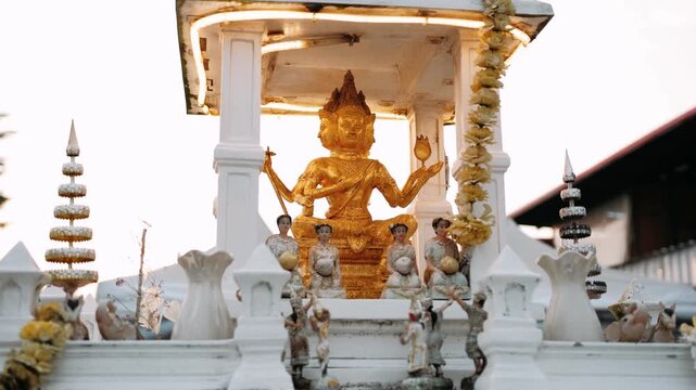 Golden brahma statue, surrounded by smaller figures and offerings, sits under an ornate white canopy in thailand.