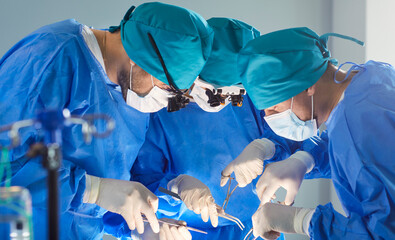 Team of professional surgeons leaning over patient during operation in surgical room. Focused medical specialists in blue scrubs and masks using surgical instruments, working together during surgery.