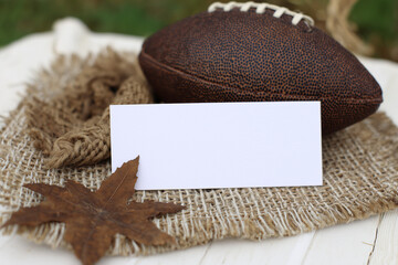 Blank white horizontal card mockup on fabric backing with rugby ball and dried maple leaves, soft natural light.