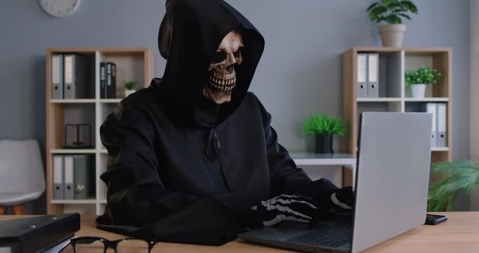 Grim Reaper of Death finding victim online, Internet scam cybercrime fraud, illegal criminal activity on laptop, spam, Halloween cosplay costume, black hooded cape, skull face mask, skeleton gloves