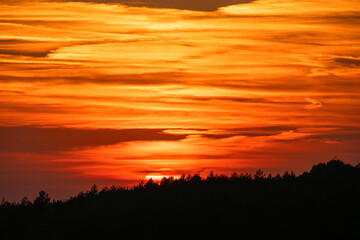 A brilliant sunset blazes behind a forested horizon, illuminating layered clouds in fiery tones of orange, red, and gold.