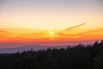 A glowing sunset bathes the forest and distant mountains in warm, tranquil light.