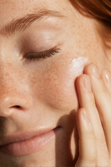 Skincare promotional image of a young woman applying cream to her face with fingers.