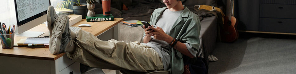 Fototapeta na wymiar Teenage Caucasian boy sitting with legs on desk using smartphone while ignoring computer screen in bedroom, demonstrating gadget addiction and lack of focus on studies