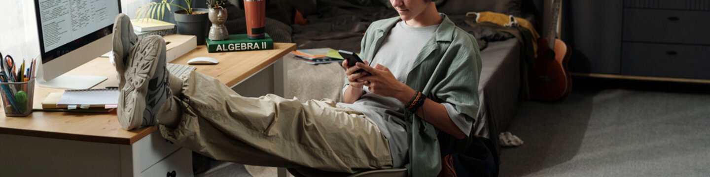 Teenage Caucasian boy sitting with legs on desk using smartphone while ignoring computer screen in bedroom, demonstrating gadget addiction and lack of focus on studies