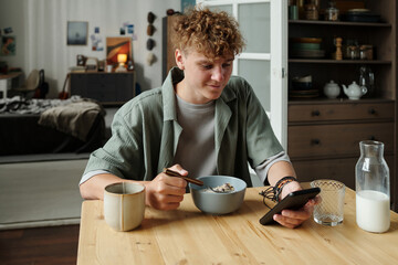 Caucasian teenage boy sitting at table eating breakfast cereal while holding smartphone in hand,...