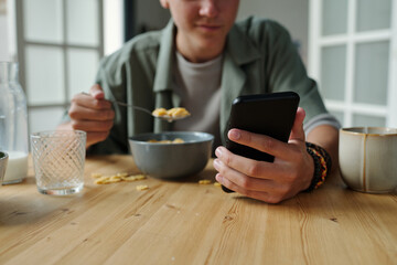 Teenager boy with light skin sitting at table holding smartphone in one hand while eating breakfast...